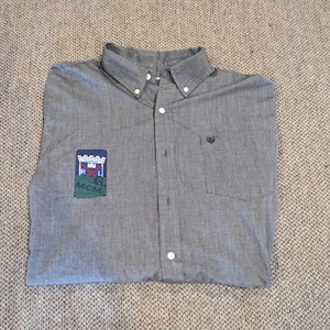 Puede incluir: Camisa gris abotonada con cuello abotonado y bolsillo en el pecho. La camisa presenta un parche con el texto "RAS MCM" y un diseño de escudo. La camisa está doblada y colocada plana.