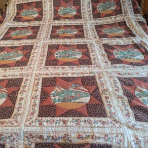 May include: A patchwork quilt with a repeating pattern of apples and stars. The quilt features squares of red, brown, and cream fabric, with floral and apple designs. The quilt is made of soft fabric and is likely for a bed.
