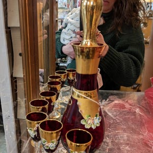 Może przedstawiać: Karafka z rubinowego szkła ze złotym korkiem i pasującymi kieliszkami do wina. Karafka i kieliszki mają złote obramowania i kwiatowe akcenty. Zestaw jest wystawiony na szklanej półce.