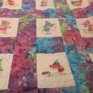 Op de afbeelding: Een handgemaakte quilt met een tie-dye patroon in tinten paars, blauw en roze. De quilt heeft geborduurde vierkanten met grillige feeënillustraties in verschillende poses, met hoeden en jurken. De quilt is een gezellig, decoratief item.