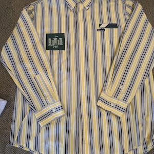 Può includere: Camicia a righe verticali gialle, blu navy e bianche. La camicia a maniche lunghe ha un colletto button-down e una tasca sul petto. Una toppa verde con un disegno di castello e il numero romano VIII è sul petto sinistro.