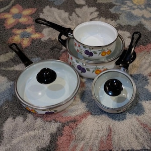 Puede incluir: Un juego de utensilios de cocina de esmalte blanco con asas y tapas negras. Las ollas y sartenes están decoradas con un colorido diseño de frutas. El juego incluye una sartén, una cacerola y dos ollas de diferentes tamaños.