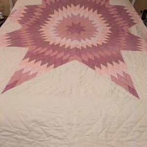 May include: A large, star-shaped quilt in shades of pink and white, centered on a bed. The quilt features a detailed geometric pattern with a central star design and a quilted, off-white background. The bed has a white headboard.
