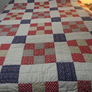 Könnte beinhalten: Eine Patchwork-Decke mit weißem Hintergrund und Quadraten aus rotem, blauem und weißem, gemustertem Stoff mit Blumenmotiven. Die Decke ist aus verschiedenen Stoffen und Mustern gefertigt und ergibt ein einzigartiges und farbenfrohes Design.