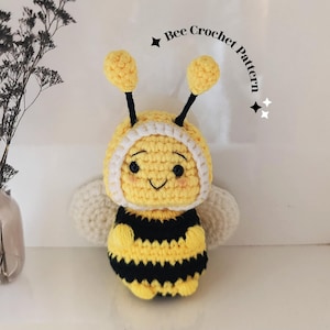 May include: A handmade crochet bee toy with a yellow body and black stripes. The bee has a smiling face, white wings, and a white-trimmed hood. The text "Bee Crochet Pattern" is above the bee.