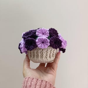 May include: A crocheted basket filled with purple and lavender flowers. The flowers are arranged in a dense cluster, with varying shades of purple. The basket is a light beige color, and the entire piece is held in a hand.