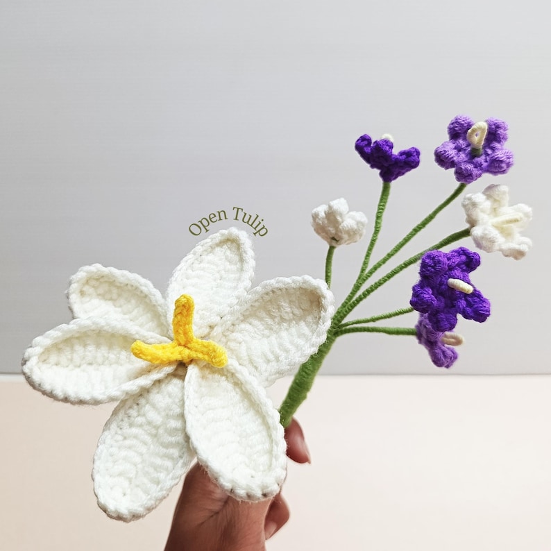 May include: A crocheted bouquet of flowers with a white open tulip and purple flowers with yellow centres. The text "Open Tulip" is written on the image.
