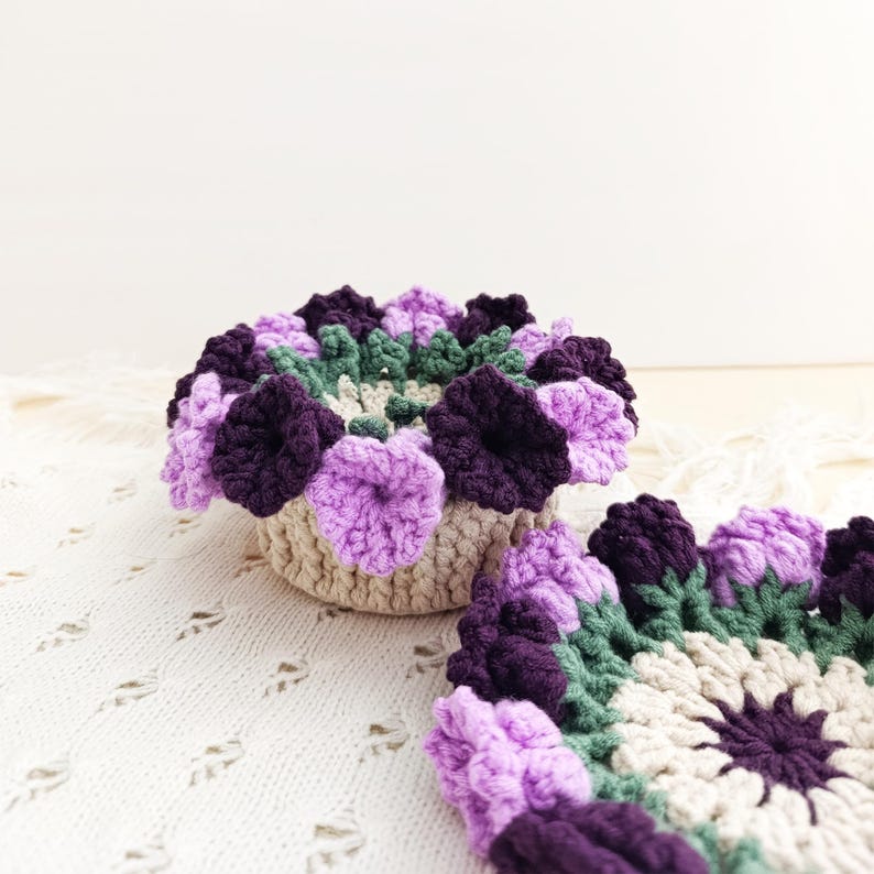 May include: A crocheted floral arrangement in a beige basket, featuring purple, lavender, and green flowers. A matching crocheted coaster is next to the basket. The handmade items are on a white, textured surface.