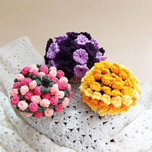 May include: Three crocheted floral spheres in shades of pink, purple, and yellow. The pink sphere has green accents, the purple sphere has darker purple accents, and the yellow sphere has lighter yellow accents. The spheres are sitting on a white crocheted blanket.