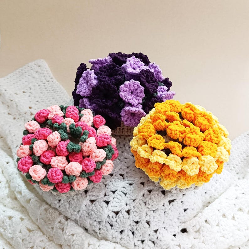 May include: Three crocheted floral spheres in shades of pink, purple, and yellow. The pink sphere has green accents, the purple sphere has darker purple accents, and the yellow sphere has lighter yellow accents. The spheres are sitting on a white crocheted blanket.