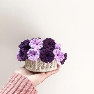 May include: A handmade crochet floral arrangement featuring purple and lavender flowers in a beige basket. The flowers are densely packed, creating a full, textured appearance. The basket is also crocheted, with a ribbed design.
