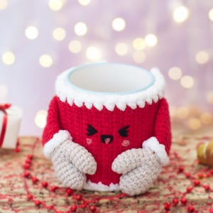 Peut inclure: Un chauffe-tasse en crochet rouge avec une bordure blanche et un visage de dessin animé mignon. Le chauffe-tasse a deux mitaines en crochet blanc sur les côtés.