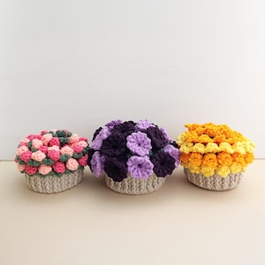 May include: Three crocheted flower arrangements in a row. The arrangements are in beige baskets. The flowers are in shades of pink, green, purple, and yellow. The flowers are arranged in a dense, rounded shape.