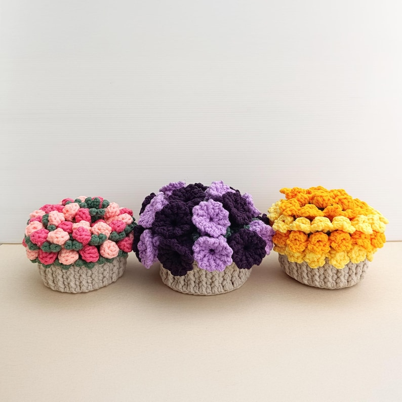 May include: Three crocheted flower arrangements in a row. The arrangements are in beige baskets. The flowers are in shades of pink, green, purple, and yellow. The flowers are arranged in a dense, rounded shape.