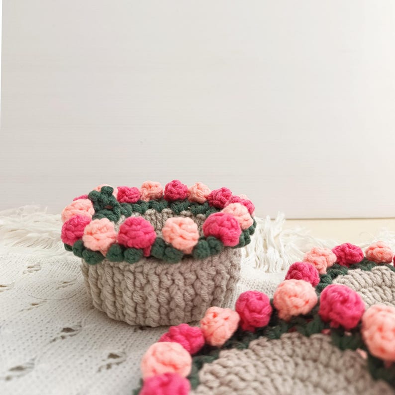 May include: A handmade, crocheted basket with a floral design. The basket is a light gray color, with a border of green leaves and pink and peach-colored flowers. A matching coaster is also visible.