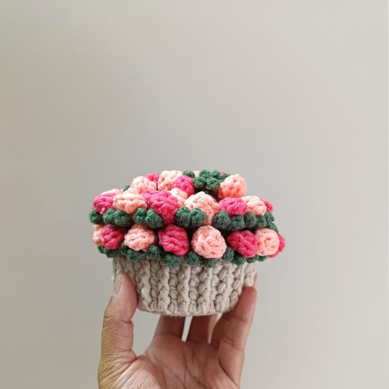 May include: A crocheted basket filled with colorful yarn flowers. The flowers are in shades of pink, peach, and green, arranged in a basket with a beige, textured design. The handmade craft is held in a hand.