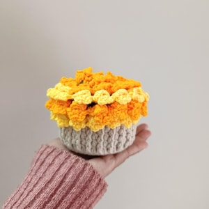 May include: A handmade crochet cupcake with a beige base and layers of yellow and orange yarn. The top is a fluffy, textured design, resembling frosting. The cupcake is held in a hand, showcasing the craft's detail and color palette.