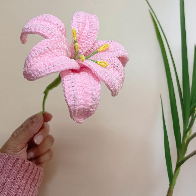May include: A pink crocheted lily flower with yellow centres. The flower is held in a hand wearing a pink sweater.