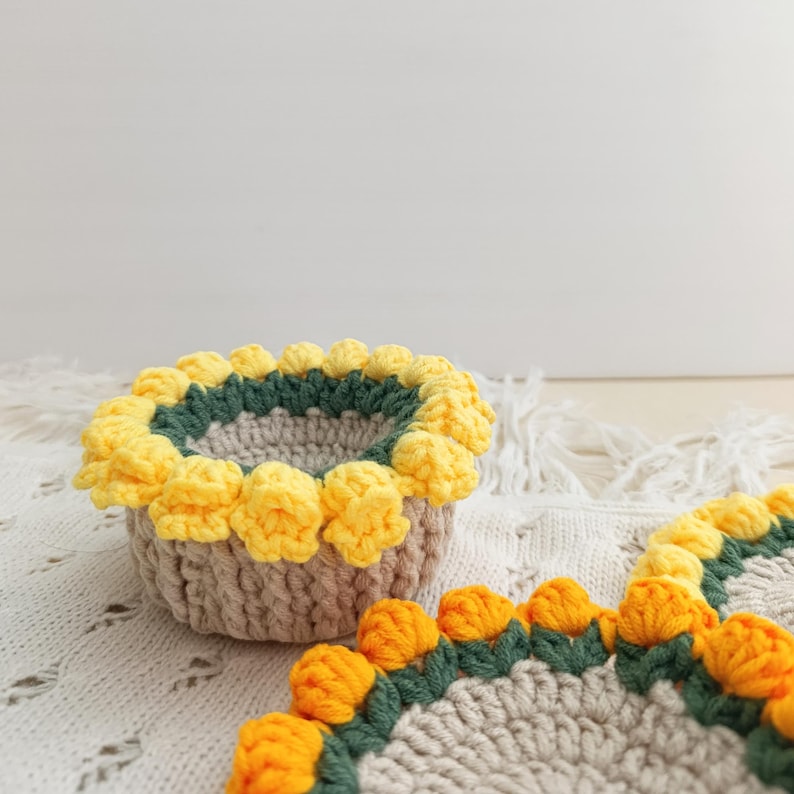 May include: A crocheted basket and coaster set. The basket is beige with a green and yellow floral trim. The coaster has a similar design. The items are handmade and sit on a white, textured surface.