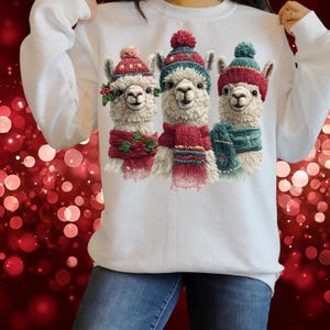 May include: White sweatshirt featuring a festive design of three llamas. Each llama wears a red and green hat and scarf. The llamas have a fluffy, textured appearance, and the overall design has a holiday theme.