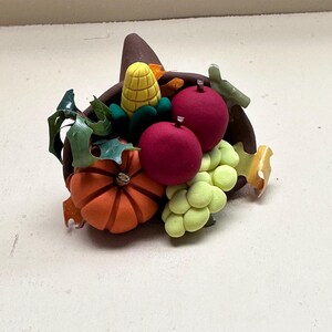 May include: A brown cornucopia filled with a harvest display featuring a bright orange pumpkin, two deep red apples, a yellow ear of corn, a cluster of light green grapes, and dark green leaves, all crafted with a smooth, matte finish.