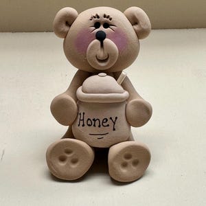 May include: A brown ceramic teddy bear figurine holding a honey pot with the word "Honey" written on it.