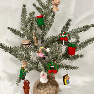 May include: A small Christmas tree decorated with handmade ornaments. The ornaments include a pig, a gift, a gingerbread man, a snowman, a wreath, and a lollipop. The tree is in a burlap sack.