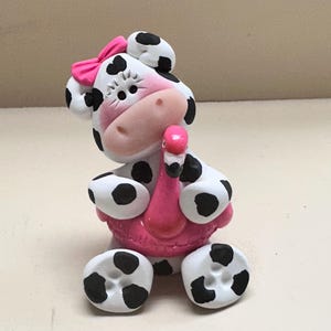 May include: A white and black spotted cow figurine with a pink bow on its head and pink cheeks. The cow is sitting down and holding a small pink flamingo pool float around its midsection. The figurine has a smooth, matte finish throughout.
