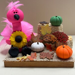 May include: A pink turkey figurine with a sunflower and black feet sits on a burlap square with fall leaves and pumpkins.