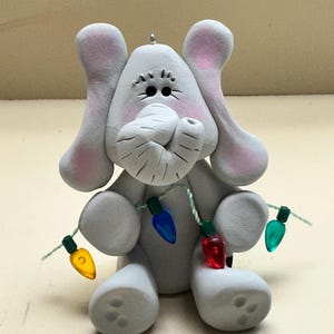 May include: A gray elephant figurine with pink-lined ears and a trunk. The elephant is decorated with a string of colorful Christmas lights. The figurine is a whimsical holiday decoration.