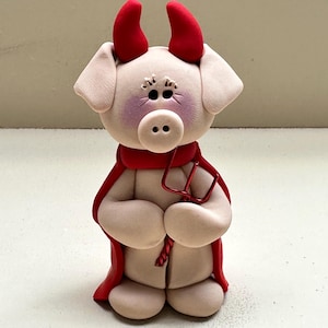 May include: A whimsical figurine of a piglet dressed as a devil. The piglet has red horns, a red cape, and a red trident. The piglet's face is light pink with darker pink accents around the eyes and snout. The figurine is made of a smooth, clay-like material.