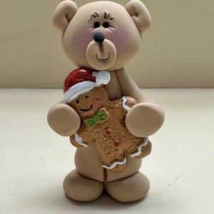 May include: A tan-colored bear figurine holding a gingerbread man. The gingerbread man wears a red and white Santa hat and a green bow tie. The bear has a black nose and eyes, with pink cheeks.