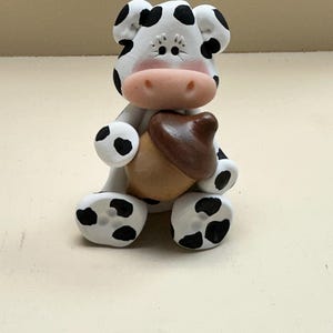 May include: A handcrafted figurine of a cow with black spots, holding a brown acorn. The cow has a pink nose and is sitting down. The figurine is white with black spots on its ears, legs, and body.