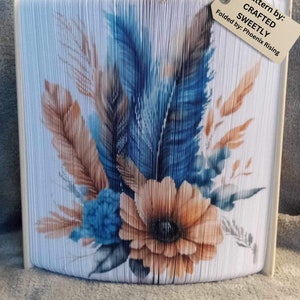May include: A white book folded into a 3D sculpture of a flower bouquet with blue and orange feathers. The book is titled "Crafted Sweetly" and the pattern is by "Phoenix Rising".