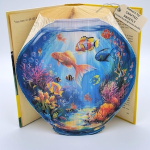 May include: A book folded into the shape of a fishbowl with colorful fish and coral reef inside. The book is open and the spine is visible. The text on the book reads "*men mest av allt till" and "Pattern by: CRAFTED SWEETLY Folded by:Brightside Studio".