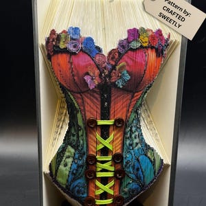 May include: A folded book art piece depicting a colorful corset design. The corset features a vibrant mix of red, orange, blue, and green hues, with brown button details and neon green lacing. The tag reads "Pattern by: CRAFTED SWEETLY."