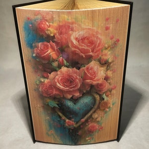 May include: A book art sculpture with a heart shape formed by the pages. The cover features a colourful image of pink roses and a blue heart.