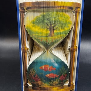 May include: A unique book art sculpture in the shape of an hourglass. The top half features a tree in a field, while the bottom half depicts fish in an aquarium. The book has a blue cover and gold accents.