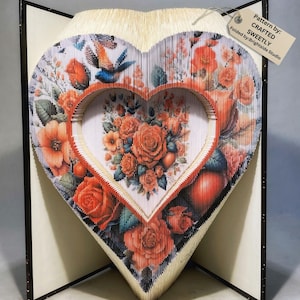 May include: A heart-shaped book sculpture made from folded pages, featuring a floral design with red roses and a blue bird. The sculpture is standing on a black book stand. The tag attached to the sculpture reads "Pattern by: CRAFTED SWEETLY Folded by: Brightside Studio".