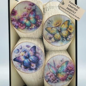 Może przedstawiać: Rzeźba książkowa przedstawiająca cztery motyle z kwiatowymi wzorami. Motyle są w odcieniach niebieskiego, fioletowego, żółtego i pomarańczowego. Książka jest złożona w trójwymiarowy kształt. Tekst na etykiecie brzmi "Pattern by: CRAFTED SWEETLY Folded by: Brightside Studio".