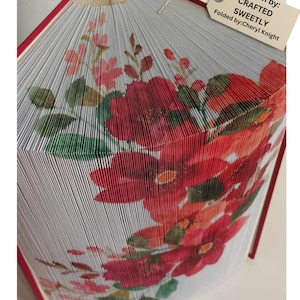 Puede incluir: Un libro doblado en una forma tridimensional, que se asemeja a un ramo de flores rojas y rosas con hojas verdes. El libro se titula "CRAFTED SWEETLY" y el patrón es de Cheryl Knight.