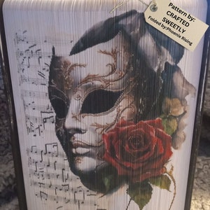 May include: A book art piece featuring a Venetian mask design with a red rose and musical notes. The artwork is folded from pages, creating a unique 3D effect. The tag reads "Pattern by: CRAFTED SWEETLY Folded by: Phoenix Rising."
