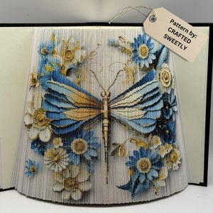 May include: A blue and yellow butterfly book fold art piece with white and blue flowers. The art piece is made from pages of a book that have been folded and glued together to create a 3D effect. The text on the tag reads "Pattern by: CRAFTED SWEETLY".