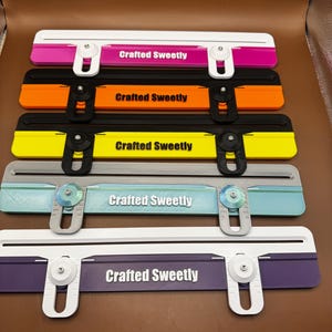 May include: A set of five horizontal paper trimmer tools, each featuring a central bar with the text Crafted Sweetly. The tools come in various color combinations including white and pink, black and orange, black and yellow, gray and teal, and white and purple, all equipped with adjustable sliding guides.
