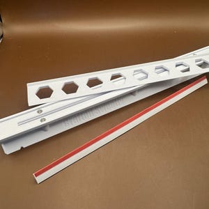 May include: A white plastic mounting rail featuring a series of hexagonal cutouts along the top edge and a flat strip with a red adhesive backing. The rail has a textured surface and includes two small silver screws embedded in the center section.
