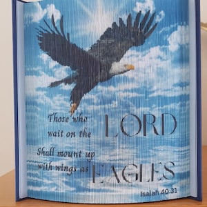 Puede incluir: Un libro azul con un diseño de águila tallada. El águila es oscura con un pico amarillo y vuela contra un cielo azul con nubes. El texto dice: "Los que esperan a Jehová tendrán nuevas fuerzas; levantarán alas como las águilas".