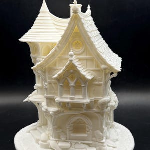 May include: A detailed, off-white miniature house with multiple stories, arched windows, and a textured roof. The house features intricate architectural details, including decorative trim and a round base. The overall design suggests a whimsical or fantasy theme.