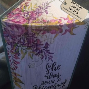 Puede incluir: Una pieza de arte de libro con un diseño floral y el texto "She was born a Wildflower". El libro está doblado en una forma tridimensional.