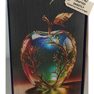 May include: A book folded into the shape of a red apple with a golden stem and leaf. The apple is filled with a colorful galaxy design. The book is titled "CRAFTED SWEETLY" and is folded by Cheryl Knight.