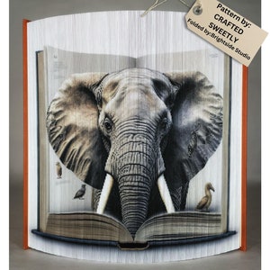 Pode incluir: Uma escultura de arte de livro de uma cabeça de elefante espiando de um livro aberto. O livro é dobrado de uma forma que cria um efeito 3D. O elefante é cinza e tem presas grandes. O livro é marrom e tem uma espinha dorsal branca. A escultura está sobre um fundo branco. Padrão por: CRAFTED SWEETLY Dobrado por: Brightside Studio